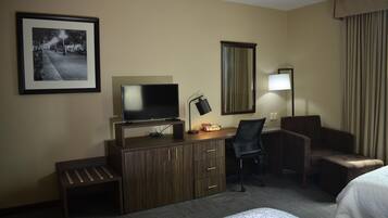 Una habitación de hotel con cama, escritorio con computadora, silla, televisor, lámpara y un cuadro en la pared.