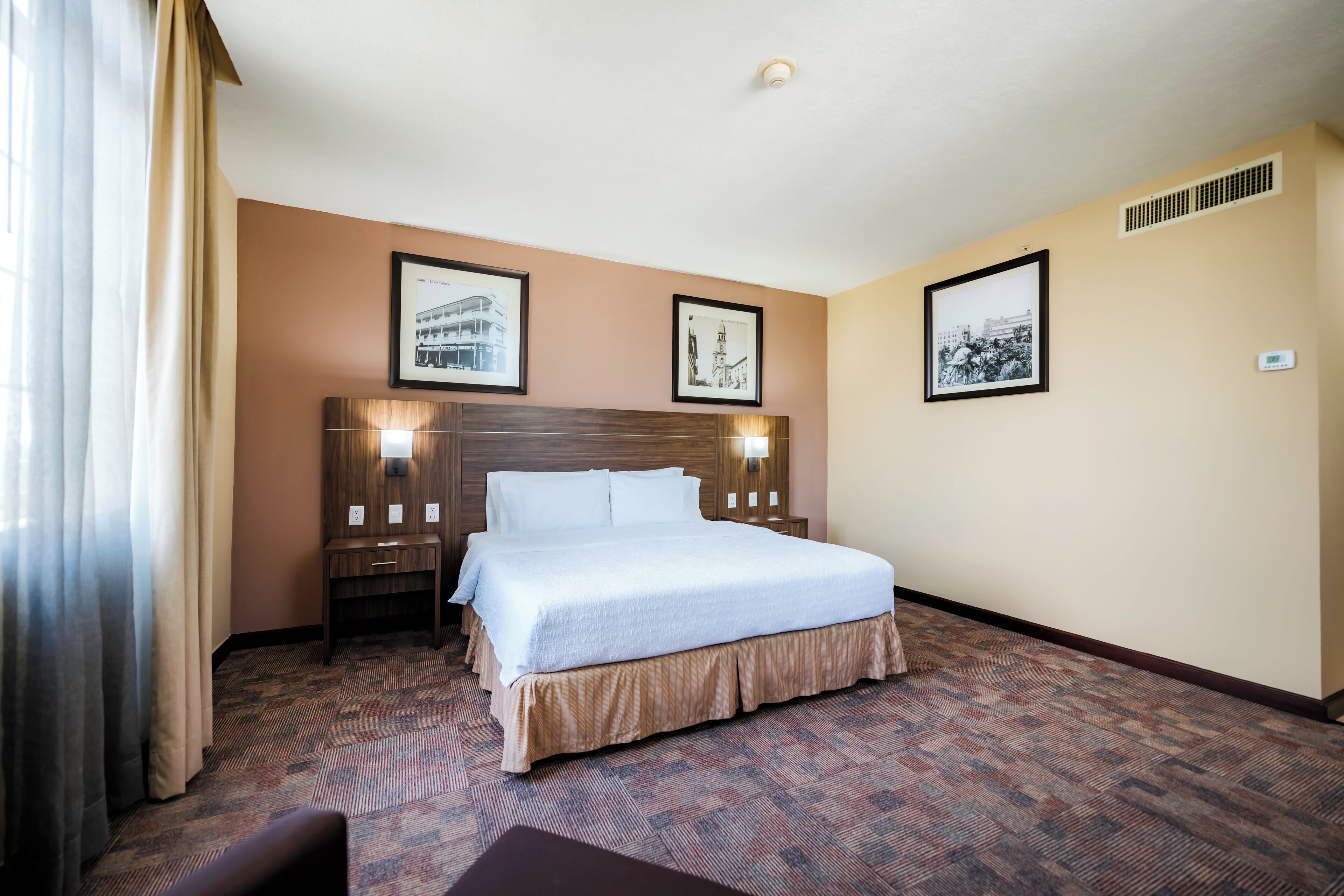 Een hotelkamer met een groot bed, een nachtkastje, een bureau en een stoel. Er hangen foto's in lijsten aan de muur en er is een raam met gordijnen.