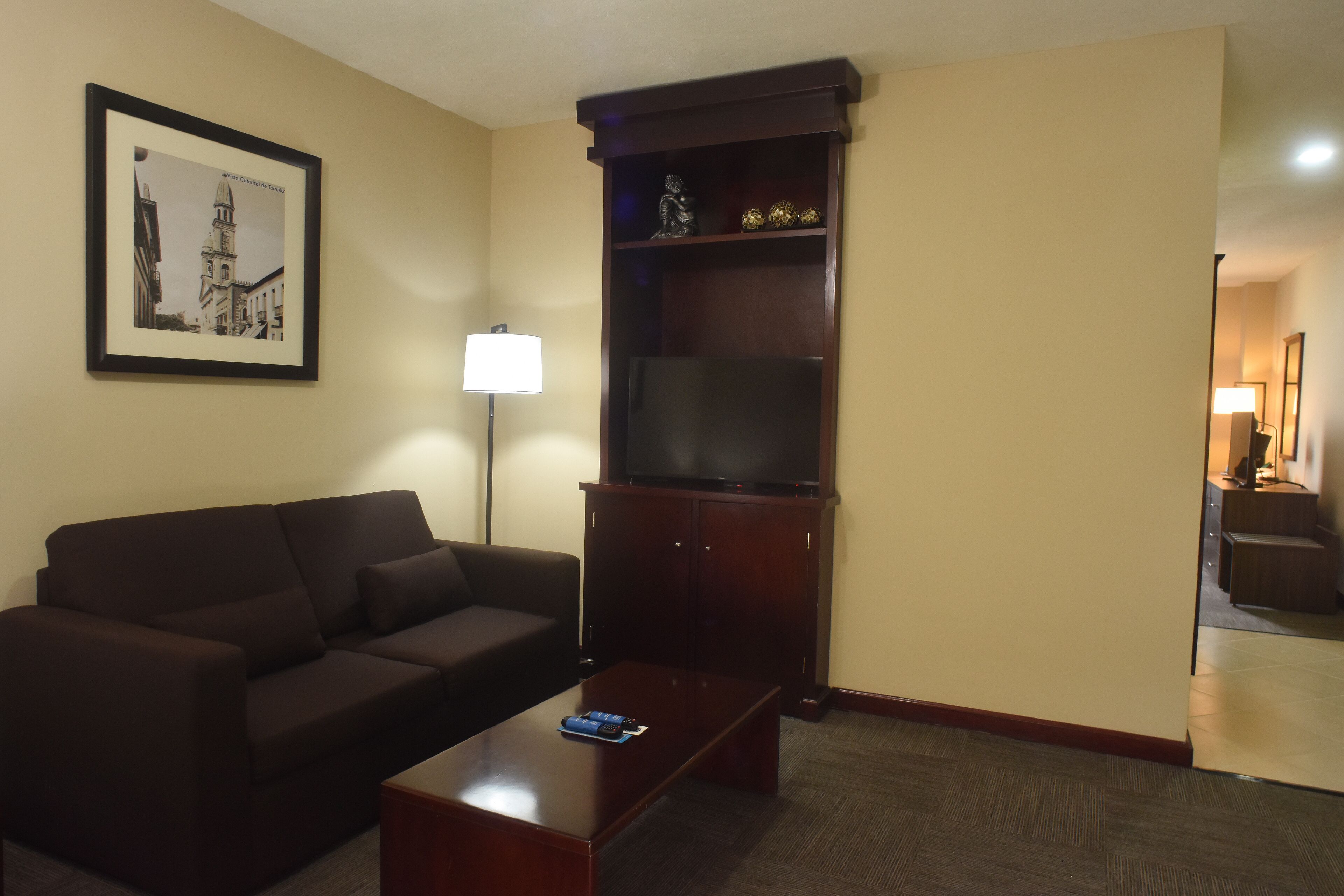 A living room with a dark brown sofa, a coffee table, a flat-screen TV on a wooden stand, a framed picture on the wall, and a lamp.