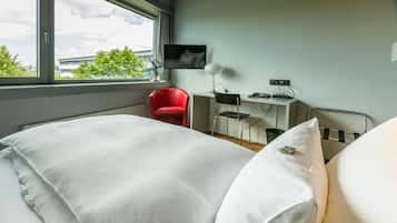 Ein Hotelzimmer mit einem großen Bett, einem Schreibtisch mit Computer, einem Stuhl, einem Fernseher und einem Fenster mit Blick auf Gebäude und Bäume.