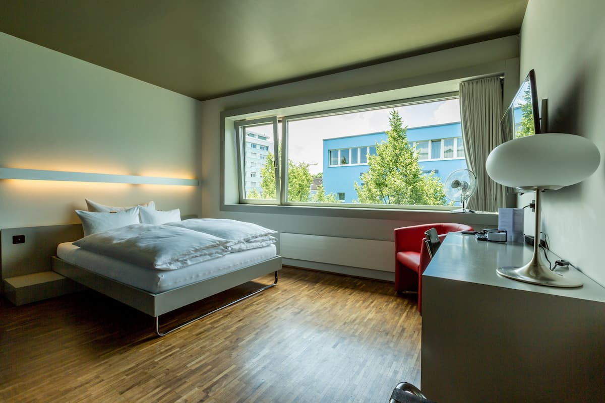 A modern bedroom with a bed, a desk, and a window with a view of buildings and trees.
