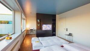 A hotel room with three beds, a desk with a laptop, a TV on the wall, and a window with a view of a building outside.