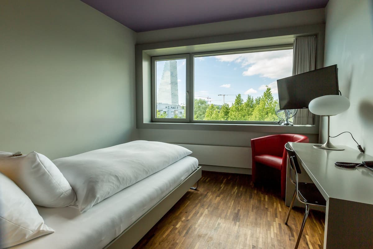A hotel room with a bed, a desk with a TV, a chair, and a window with a view of a building and trees.