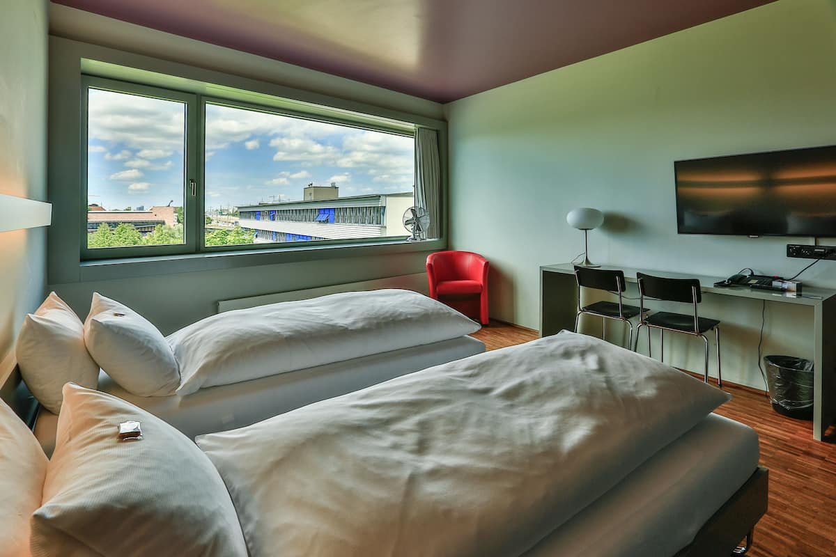 A modern hotel room with two beds, a desk, a TV, and a window with a city view.