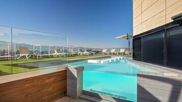 Una piscina en la azotea con paredes de vidrio transparente, una terraza de madera y sillones de descanso.