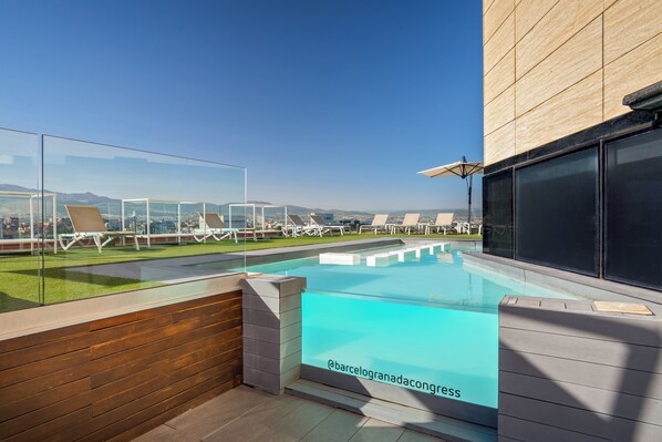 A rooftop pool with clear glass walls, a wooden deck, and lounge chairs.