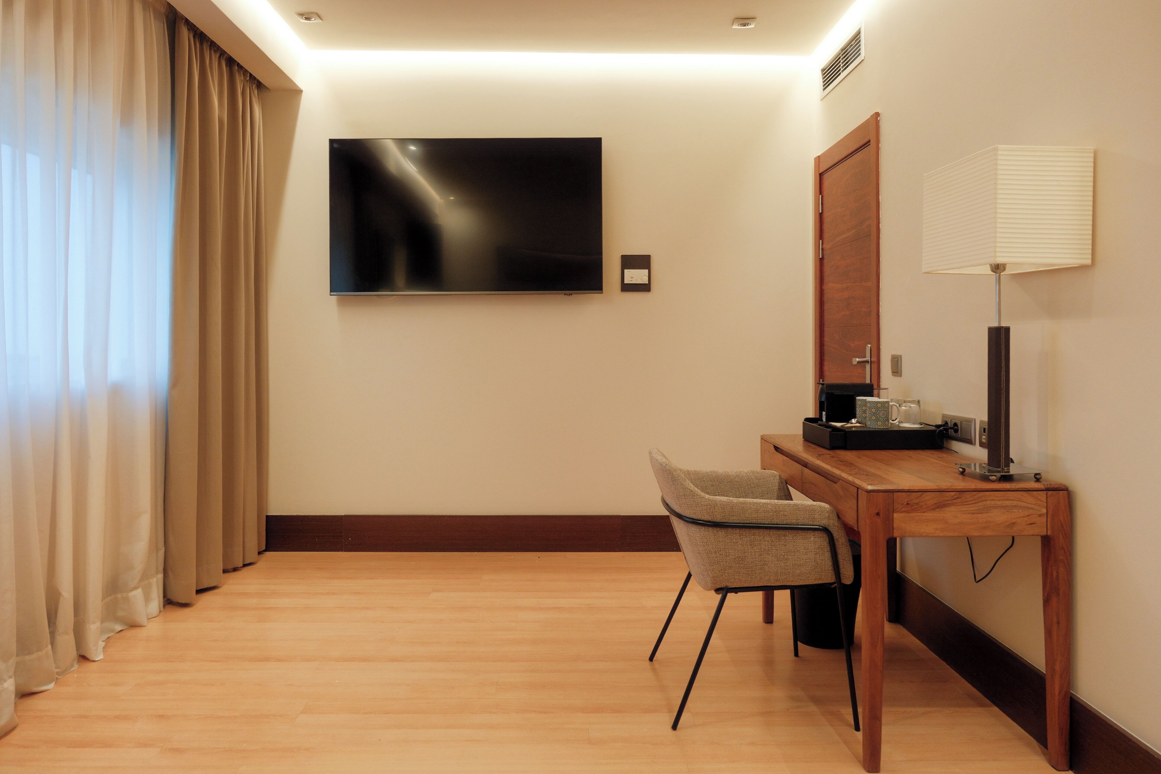 Een hotelkamer met een houten bureau, een stoel, een aan de muur bevestigde televisie en een raam met gordijnen.