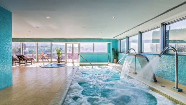 Una gran piscina interior con cascada, rodeada de sillones de descanso y vistas a la ciudad.