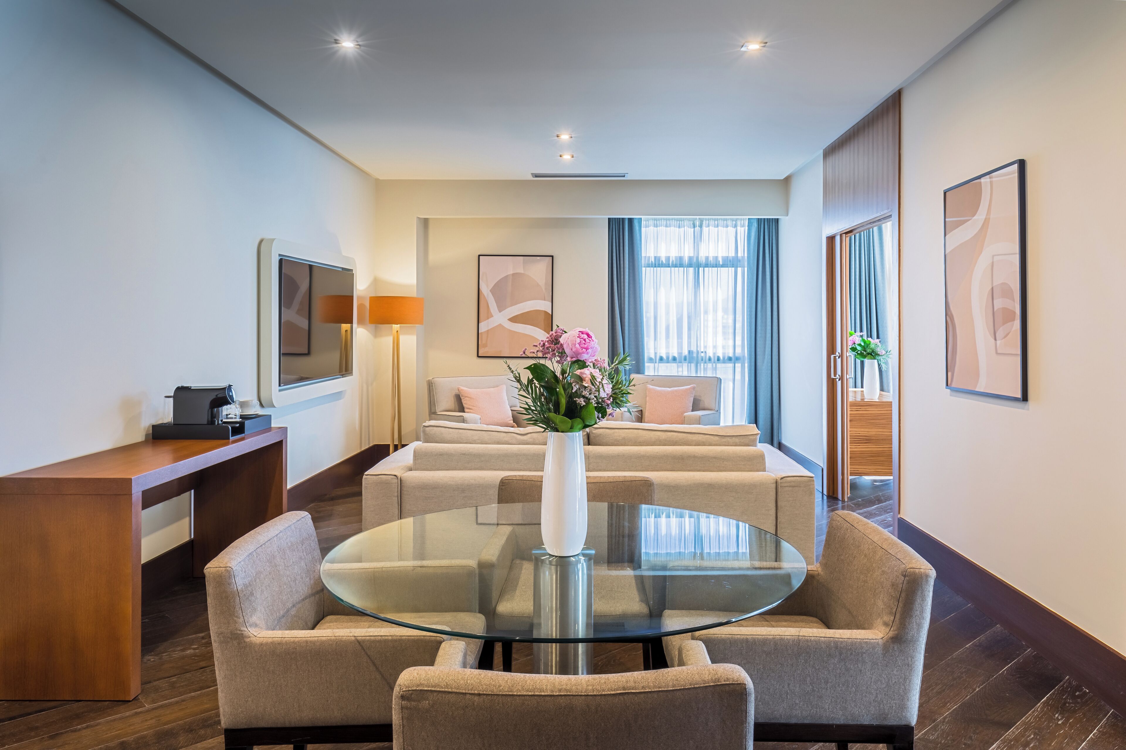 Une chambre d’hôtel moderne avec une table à manger en verre, des canapés beiges, une machine à café et des œuvres d’art accrochées aux murs.