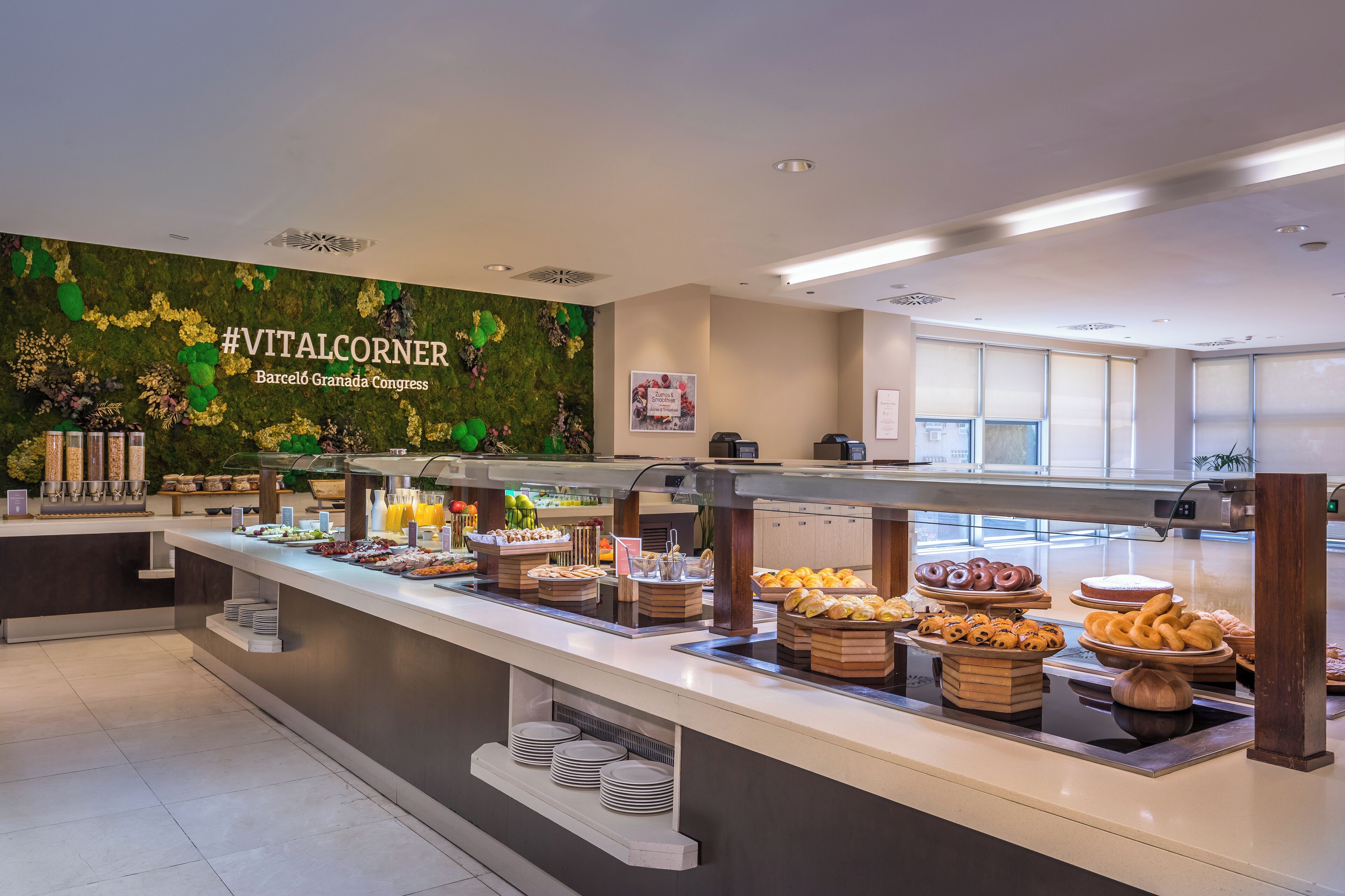 Zona de bufé con diversas opciones de comida, un cartel que dice "#VITALCORNER Barcelona Granada Congress" y una pared verde decorada con flores.