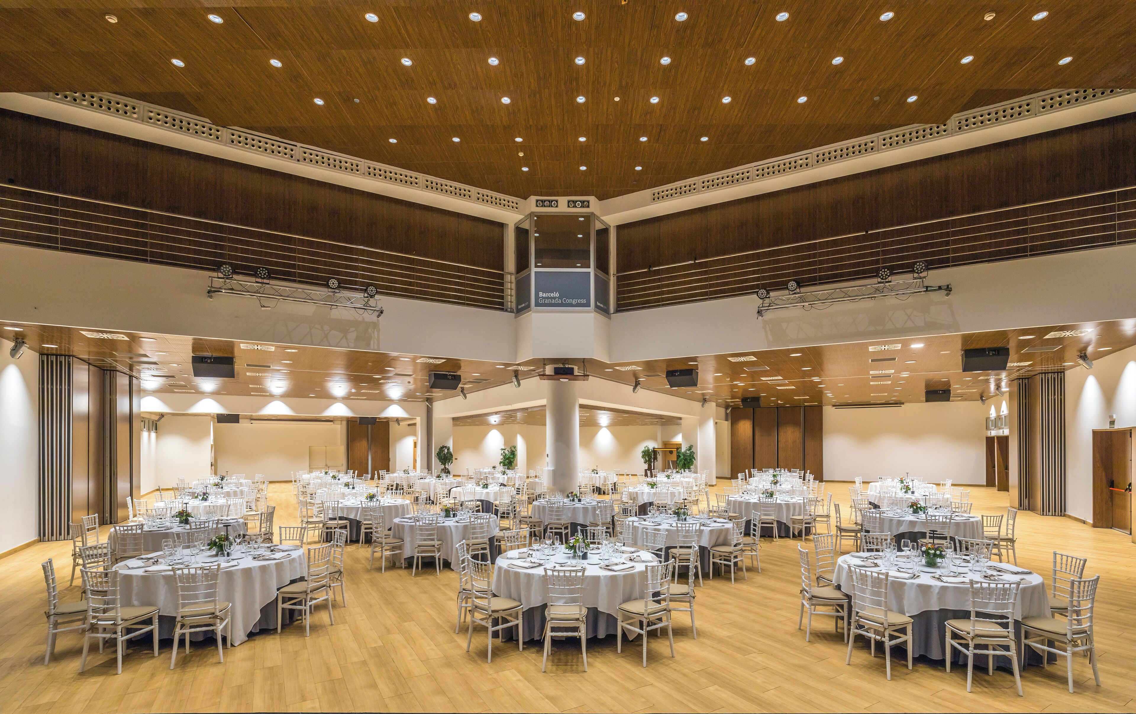 A spacious banquet hall with multiple round tables set for dining, a wooden ceiling with recessed lighting, and a stage area for presentations.