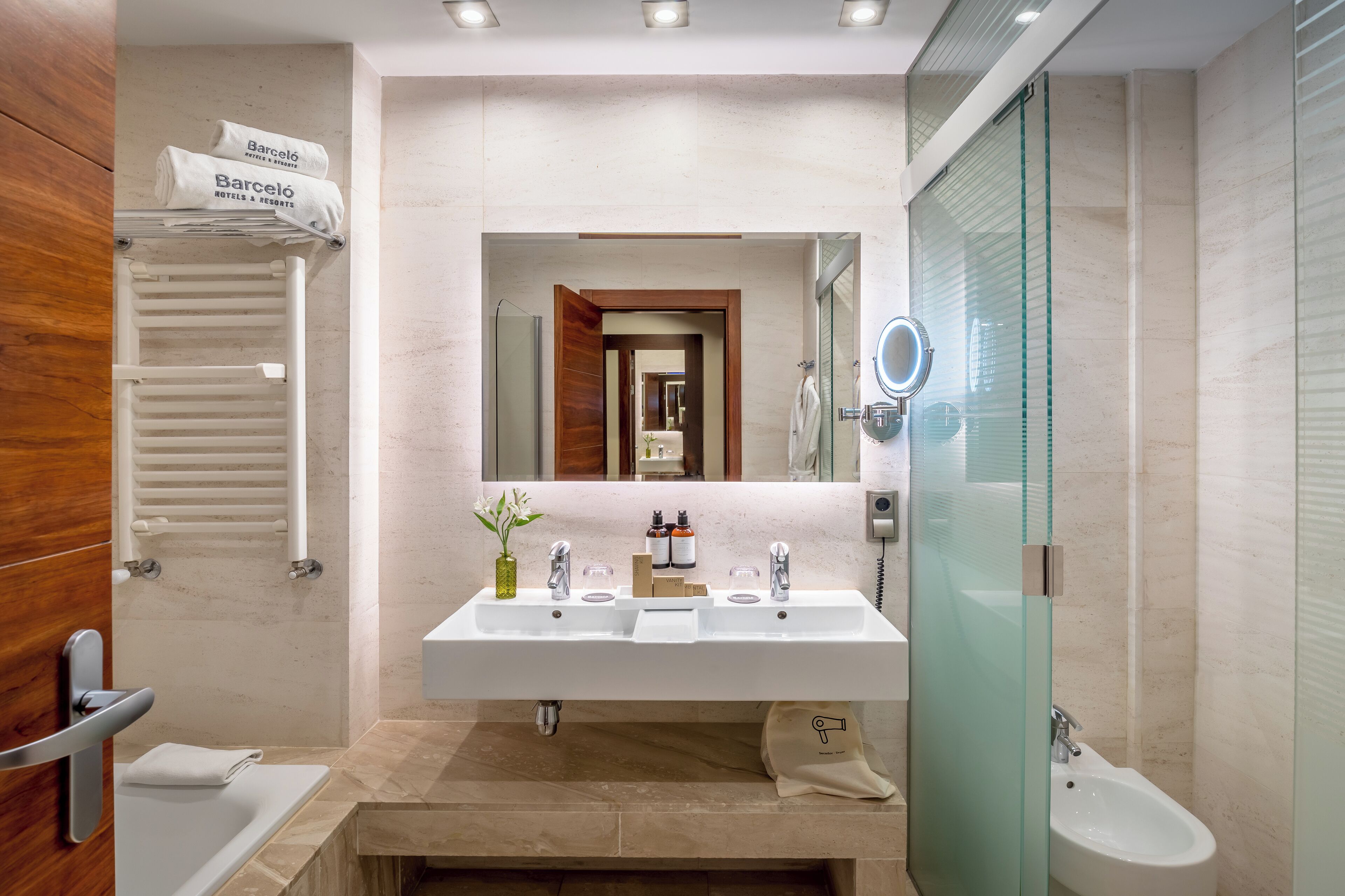 A modern bathroom with a double sink, a mirror, a glass shower enclosure, and a towel rack with a hotel name towel.