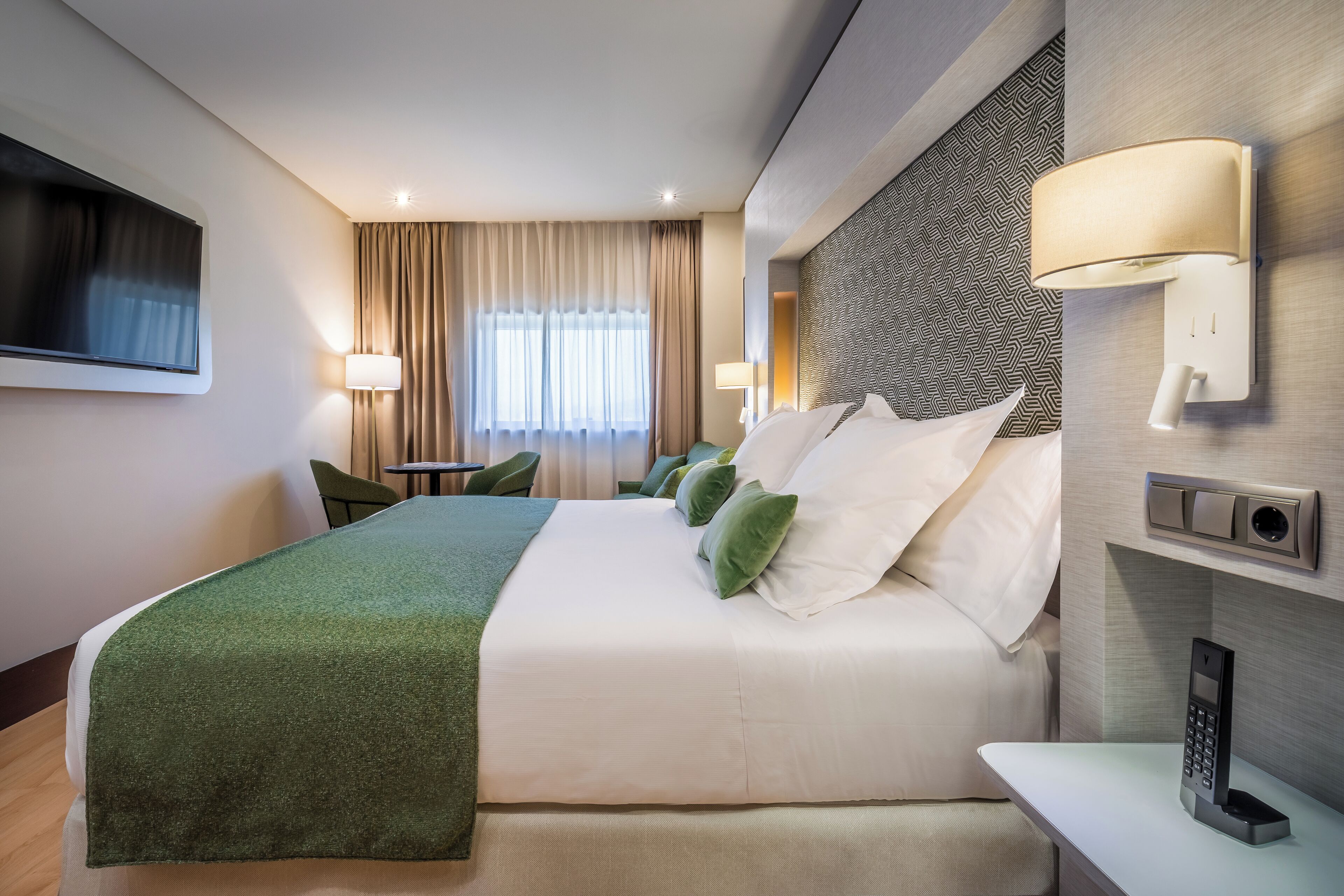 Habitación de hotel con una cama grande, un televisor montado en la pared, una mesita de noche con lámpara y un teléfono también en la pared.