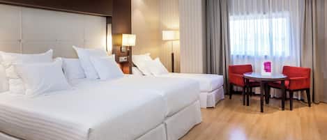 Una habitación de hotel con una cama grande, dos mesitas de noche con lámparas, una pequeña mesa con dos sillas rojas y un ventanal con cortinas.