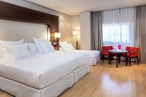 A hotel room with a large bed, two bedside tables with lamps, a small table with two red chairs, and a large window with curtains.