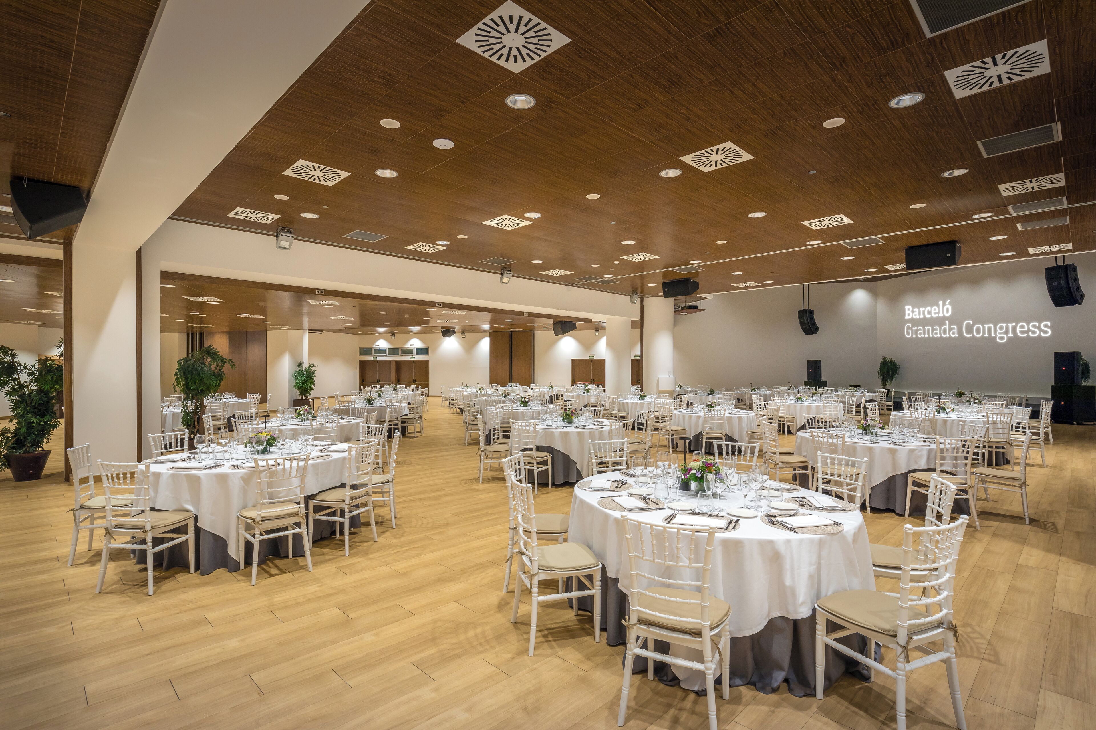 Une salle de conférence spacieuse, dotée de tables rondes et de chaises blanches, d’un plancher en bois et d’un plafond en bois orné de motifs décoratifs.