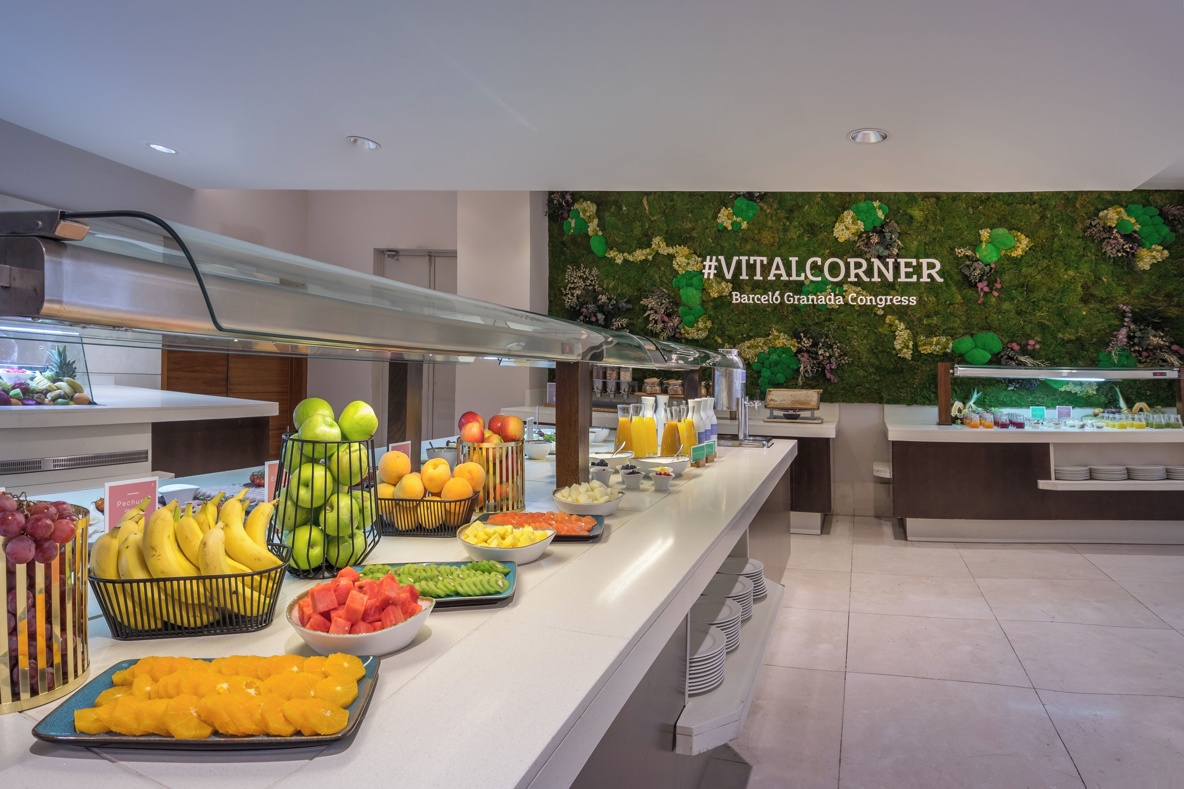 Un bufé con diversas frutas y bebidas, una barandilla de vidrio y una pared con un diseño floral y texto.