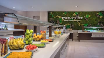 Un bufé con diversas frutas y bebidas, una barandilla de vidrio y una pared con un diseño floral y texto.