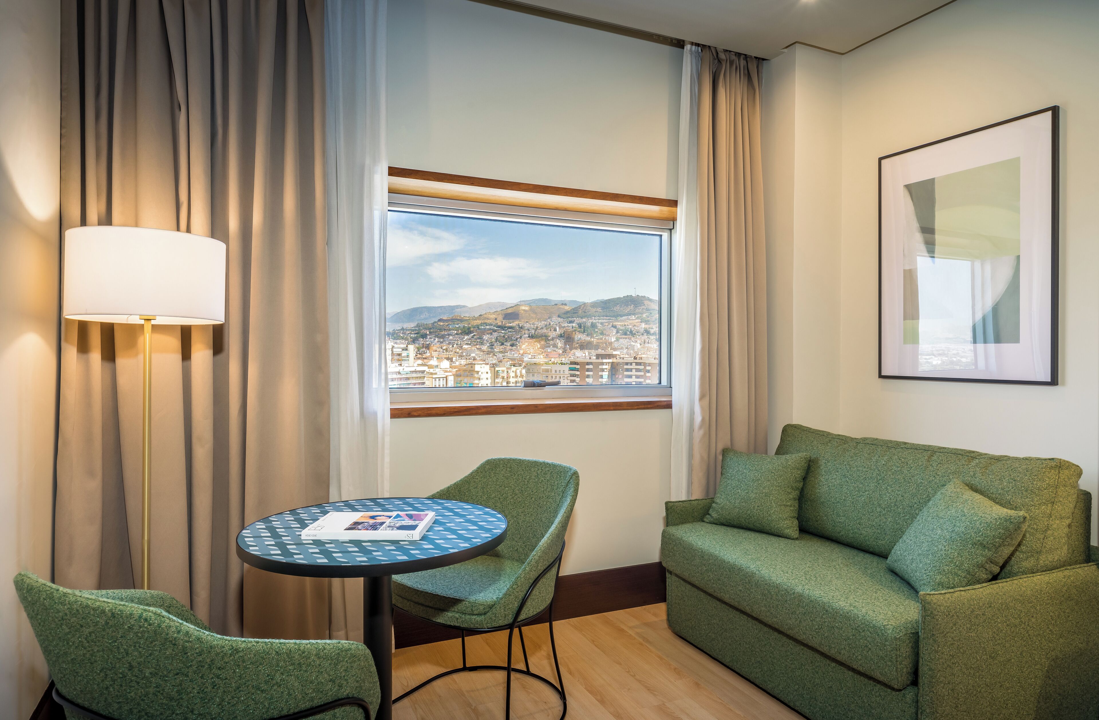 A hotel room with a view of a cityscape, a green sofa, a round table with a magazine, and a floor lamp.