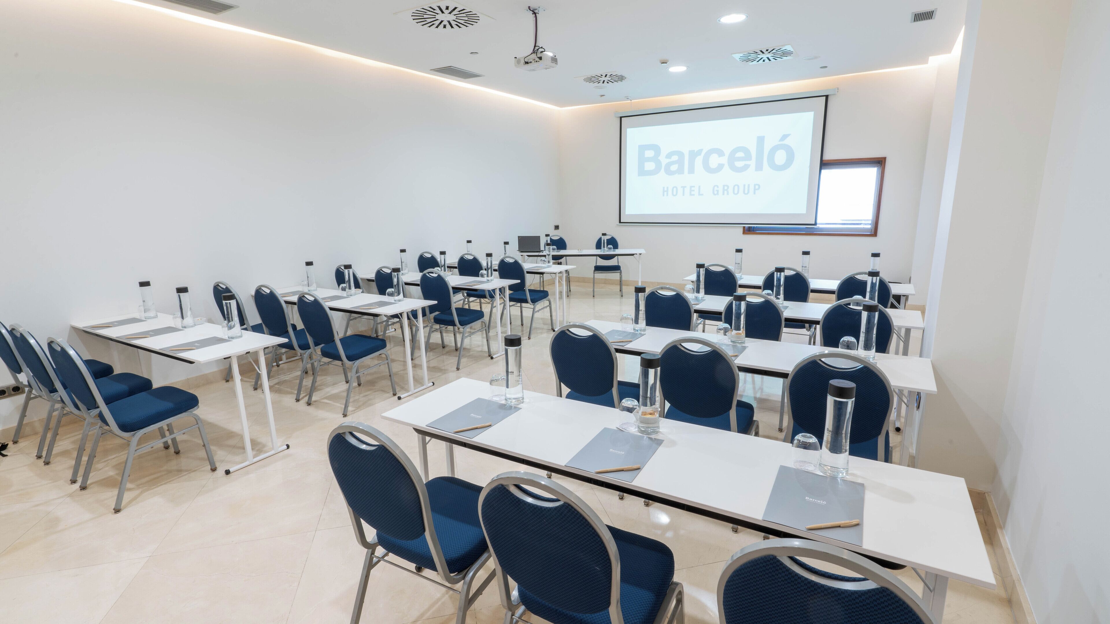 Un salón de conferencias con mesas, sillas y una pantalla de proyector que muestra 'Barceló HOTEL GROUP'.