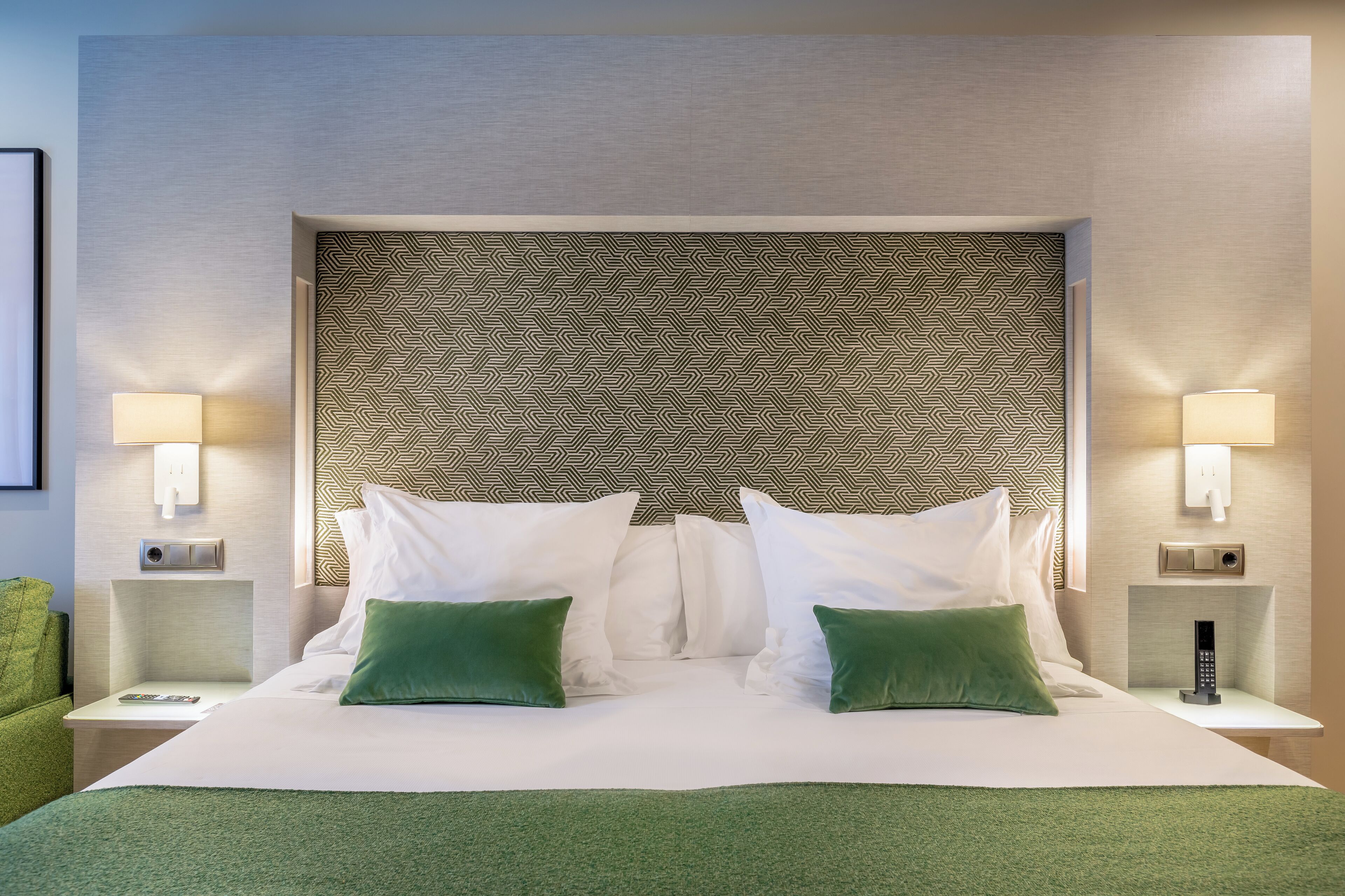 Una cama bien hecha con sábanas blancas y almohadas verdes, un cabecero estampado y lámparas de noche.