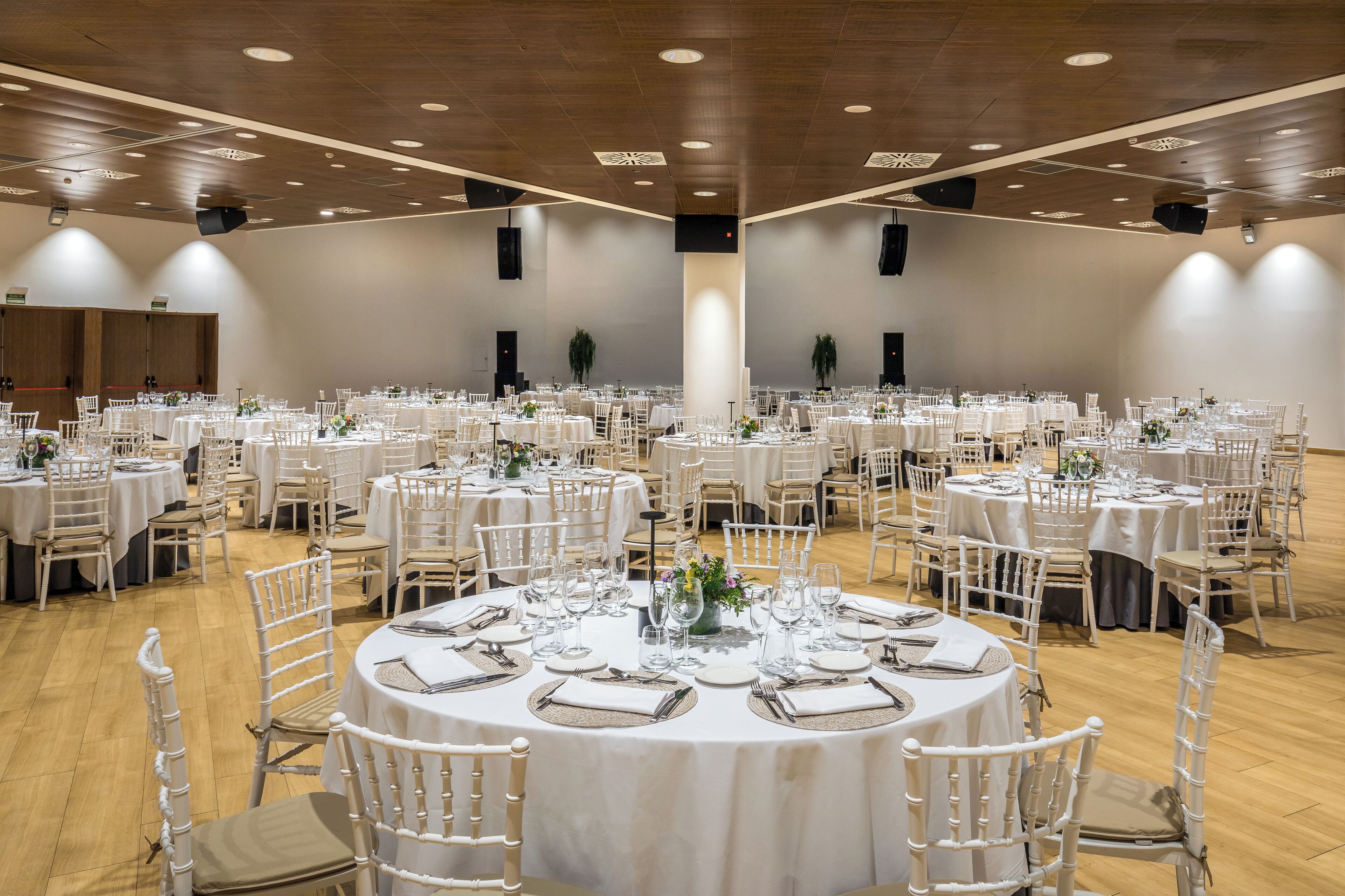 A large banquet hall with round tables set for dining, white chairs, and a wooden floor.