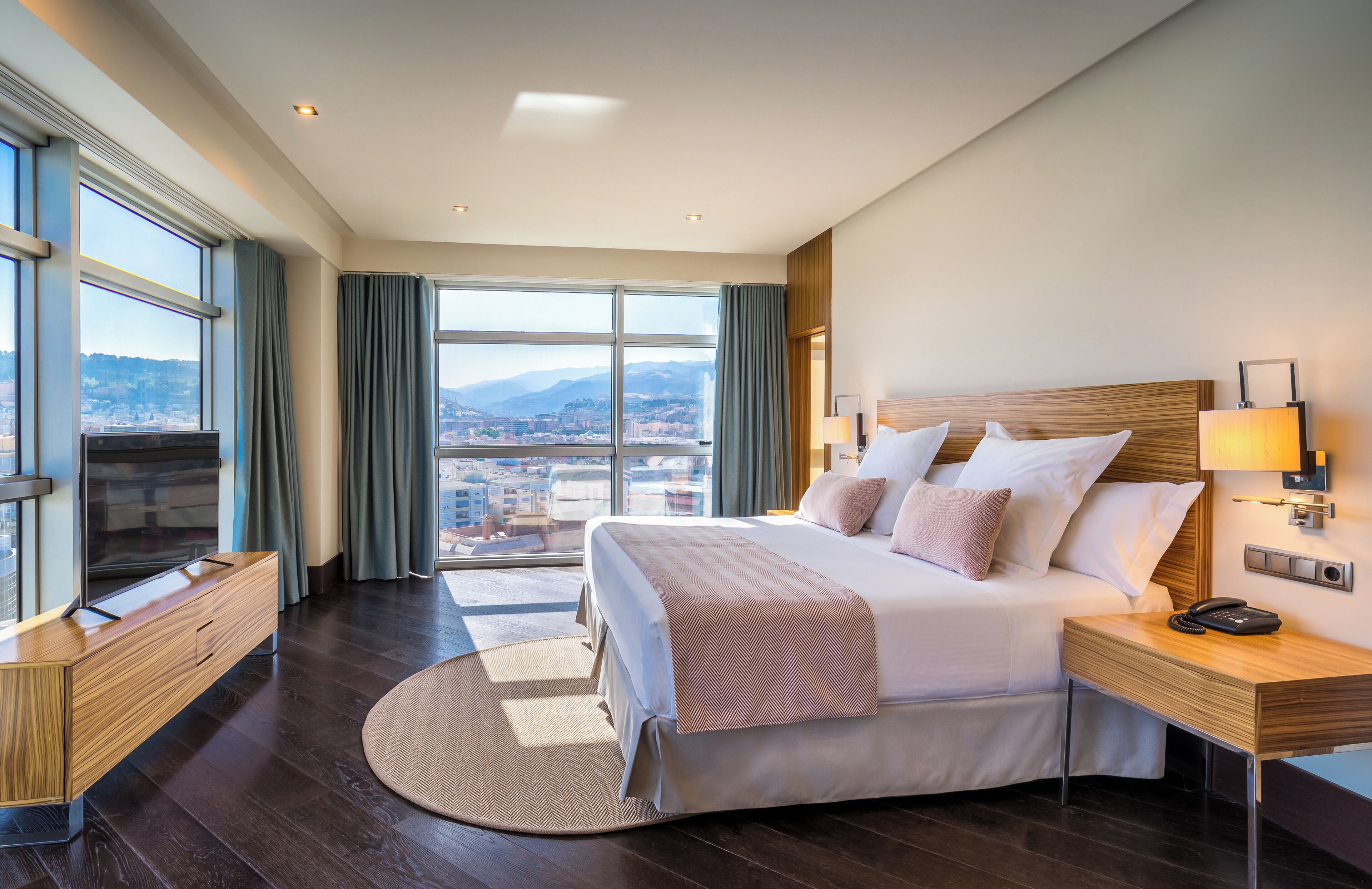 A modern hotel room with a large bed, a desk, and a view of a cityscape through large windows.