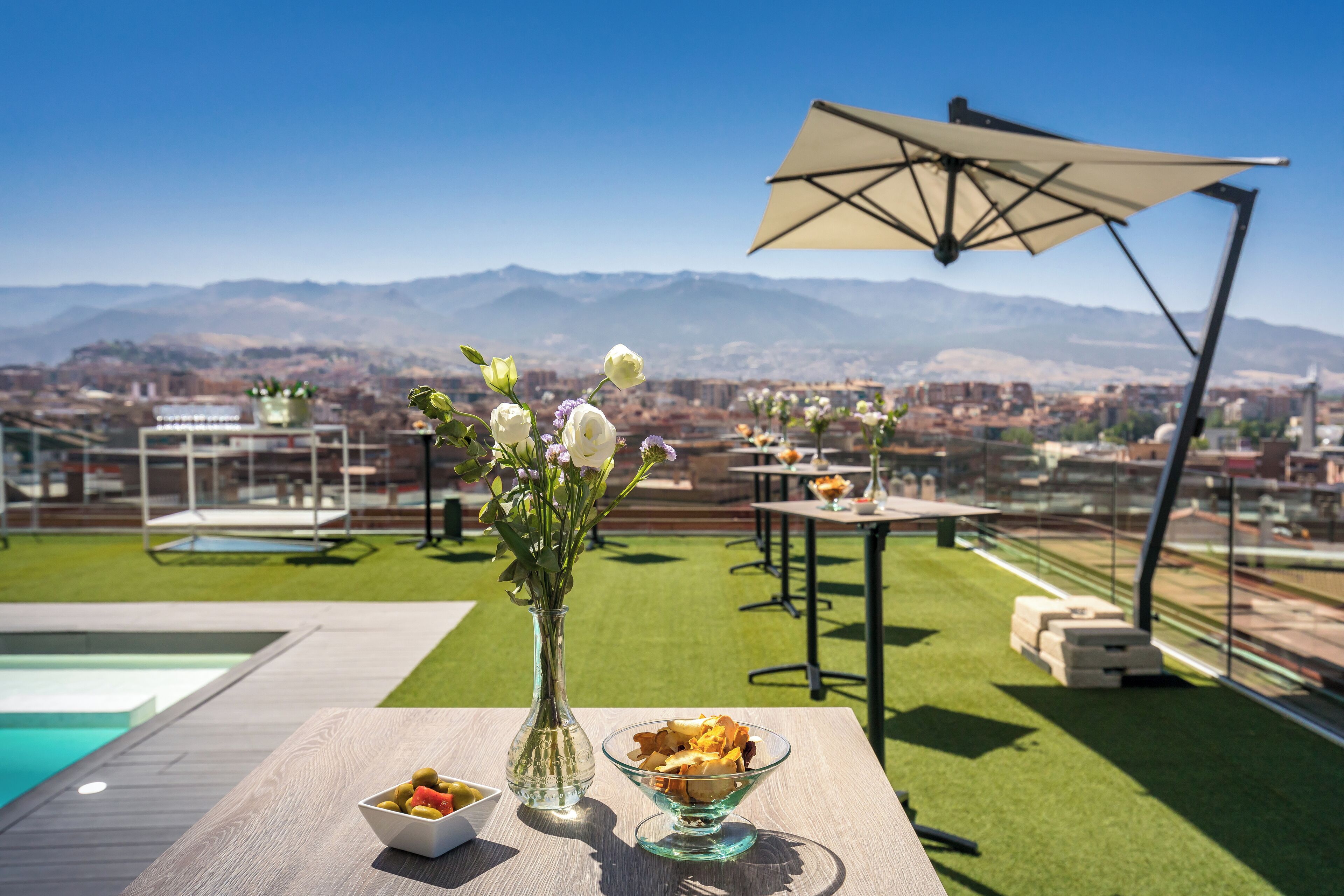 Una terraza en la azotea con piscina, una mesa de comedor con un bol de aperitivos, un jarrón de flores y un paraguas grande.