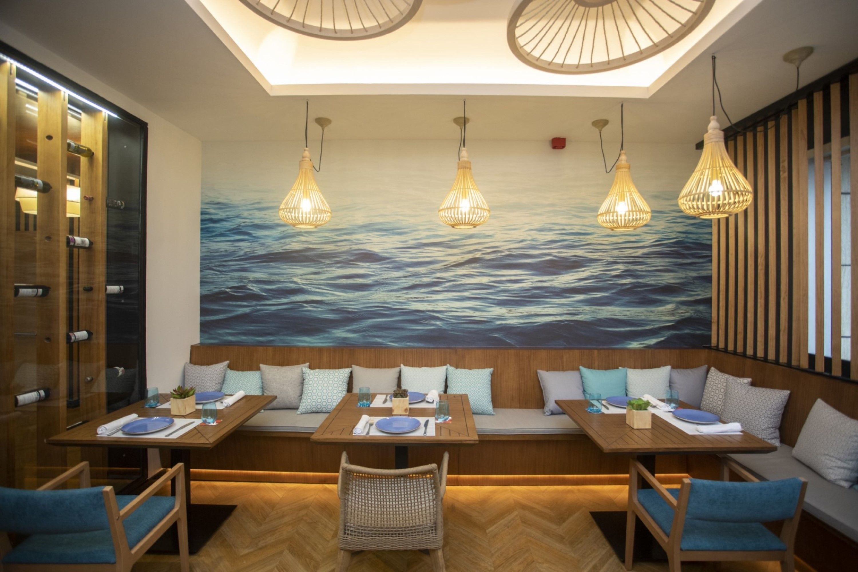 A restaurant with wooden tables and benches, hanging light fixtures, and a large ocean mural on the wall.