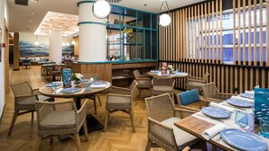 Interior de restaurante con mesas redondas, sillas de mimbre y lámparas colgantes.