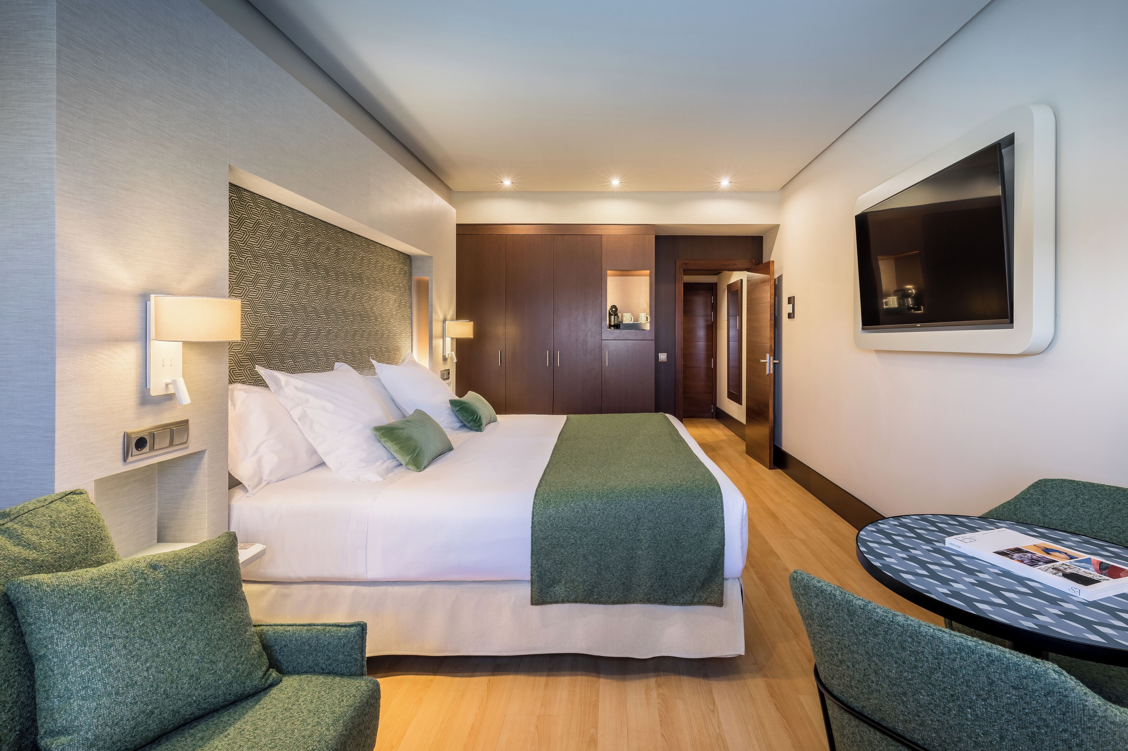 A hotel room with a large bed, a desk with a chair, a TV mounted on the wall, and a small table with a magazine.