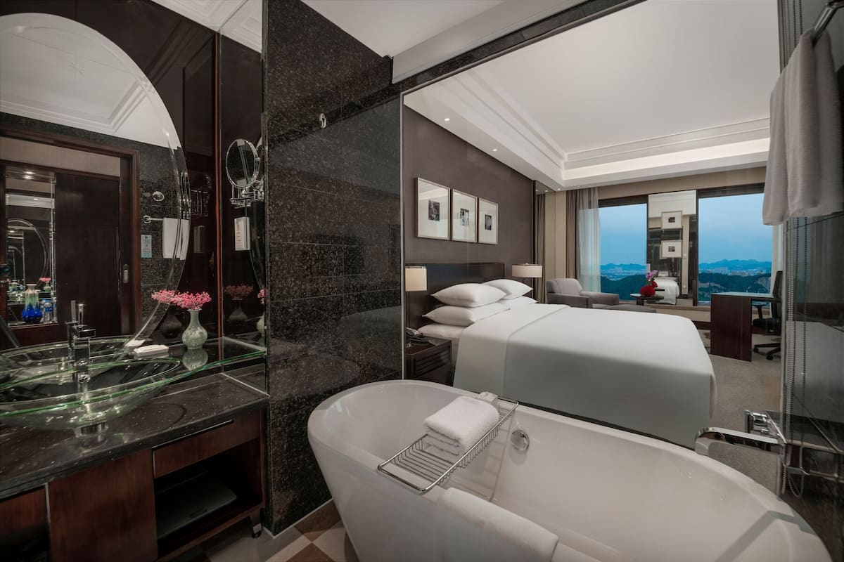 Habitación de hotel moderna con una bañera grande, un tocador con encimera de vidrio, una cama con ropa blanca y vistas a la ciudad a través de ventanas amplias.