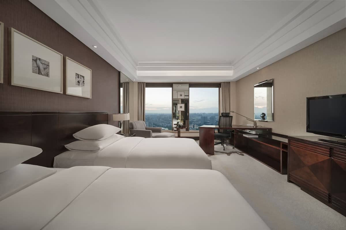 Habitación de hotel con dos camas grandes, un escritorio, televisión y vistas a la ciudad.