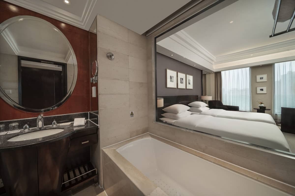 Un baño de hotel moderno con un espejo grande, una bañera, un lavamanos y una cama con vista a la ciudad.