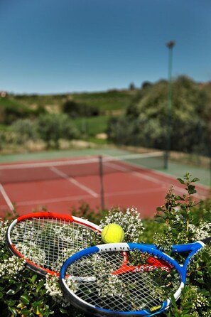 Dos raquetas de tenis sobre una cancha roja, con una pelota de tenis amarilla en medio, rodeado de follaje.