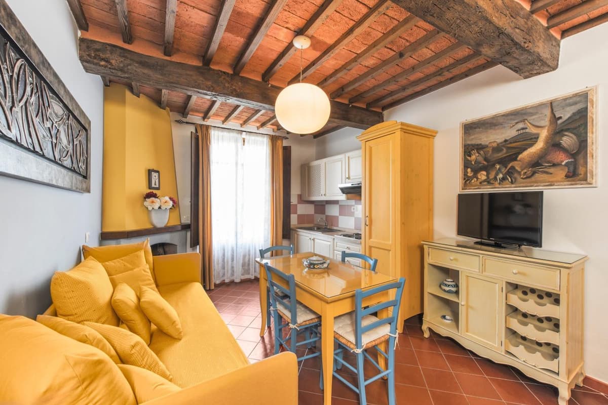 Una sala de estar acogedora con un sofá amarillo, una mesa de comedor de madera con sillas, una zona de cocina y un televisor sobre un mueble.
