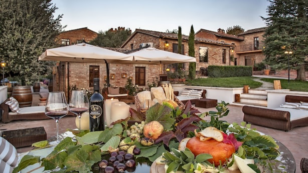 Uma área de refeições ao ar livre com mesa posta para servir alimentos e vinho, cercada por assentos externos e uma casa grande ao fundo.