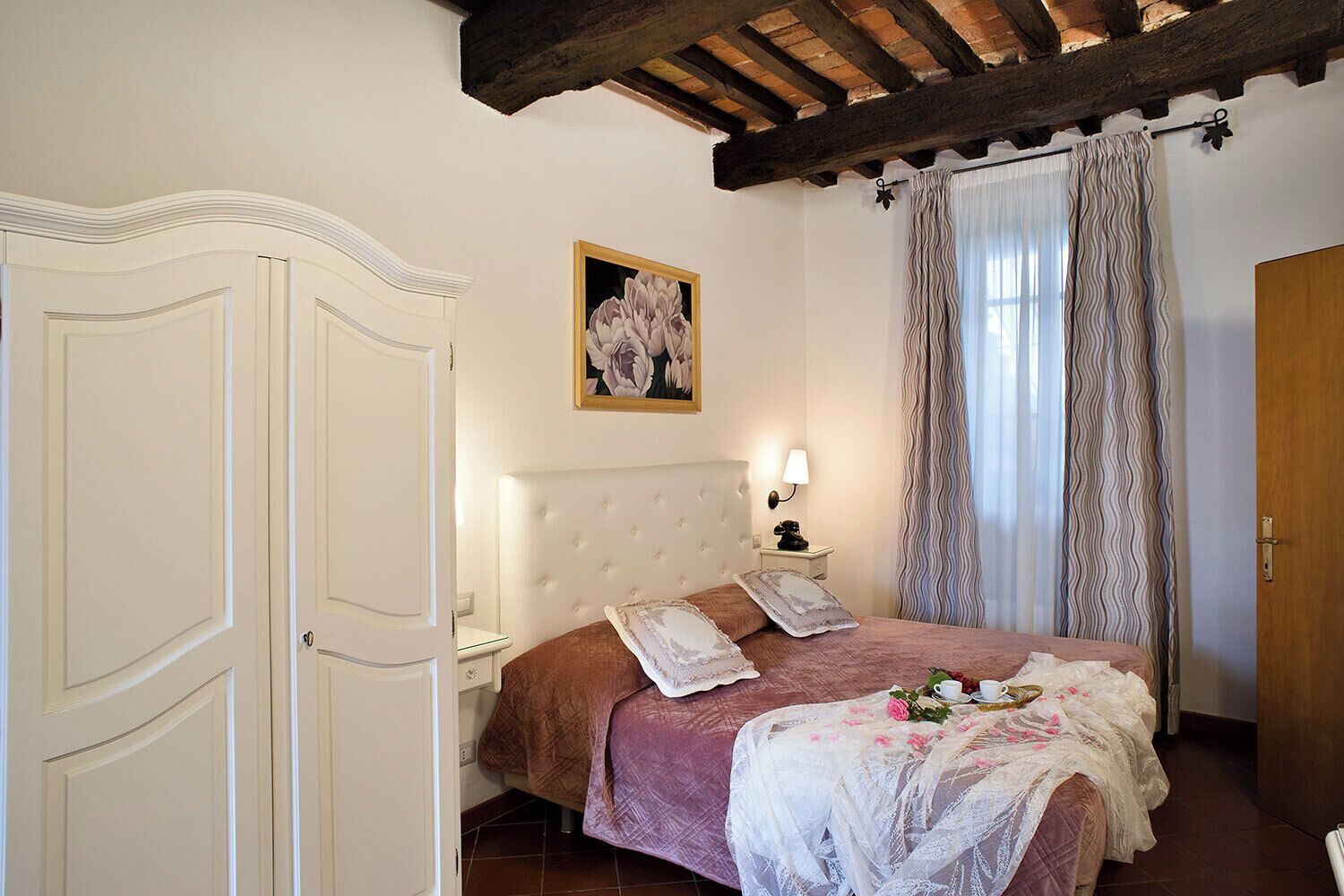 A bedroom with a large bed, a nightstand, a wall-mounted lamp, a painting, and a wooden door.