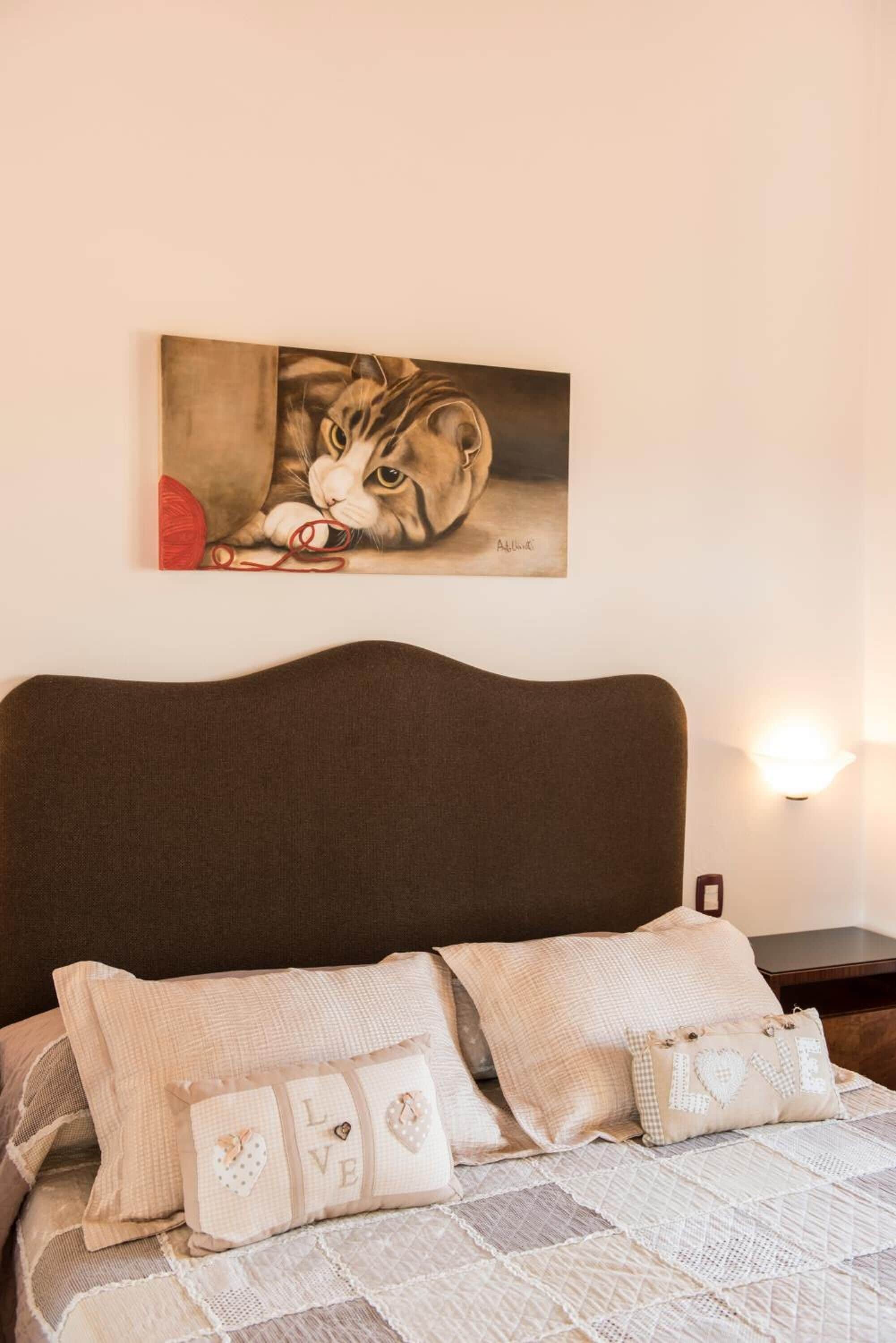 Une chambre à coucher avec un lit, des oreillers, un tableau représentant un chat accroché au mur et une table de chevet avec une lampe.