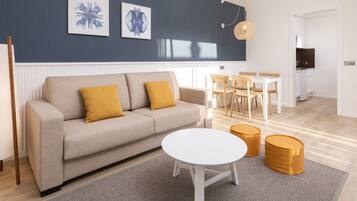 A modern living room with a sofa, a white coffee table, and two ottomans. There is a dining area with a table and chairs in the background.