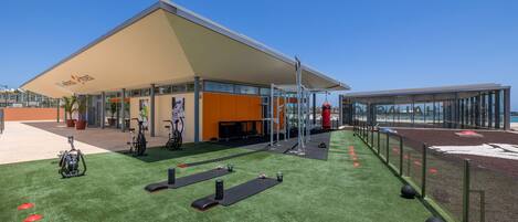 Un área de gimnasio al aire libre con equipo de ejercicio, una zona de estar cubierta y cielo despejado.