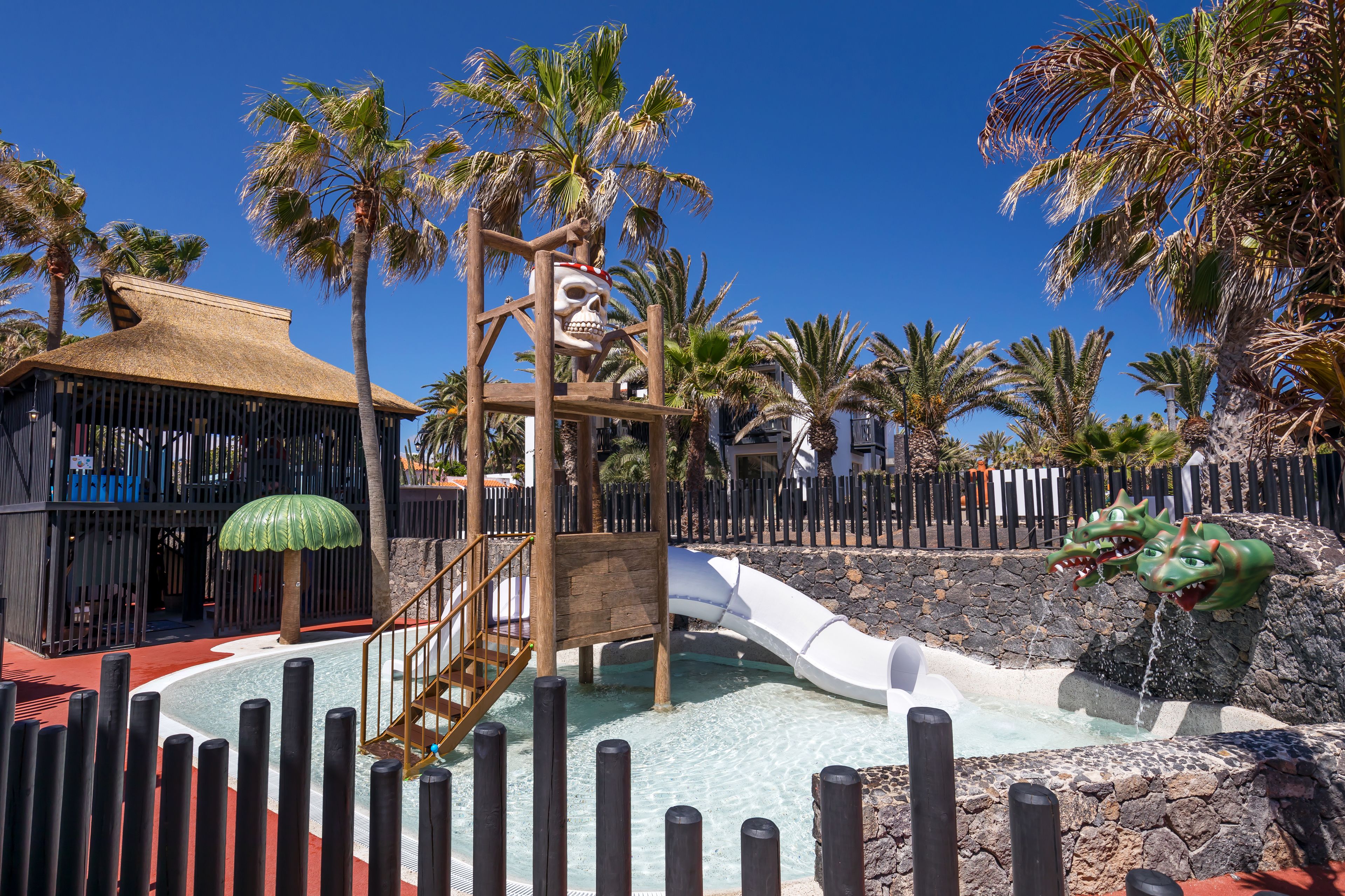 Parque acuático con tobogán, palmeras y una estructura de madera decorada con calavera.