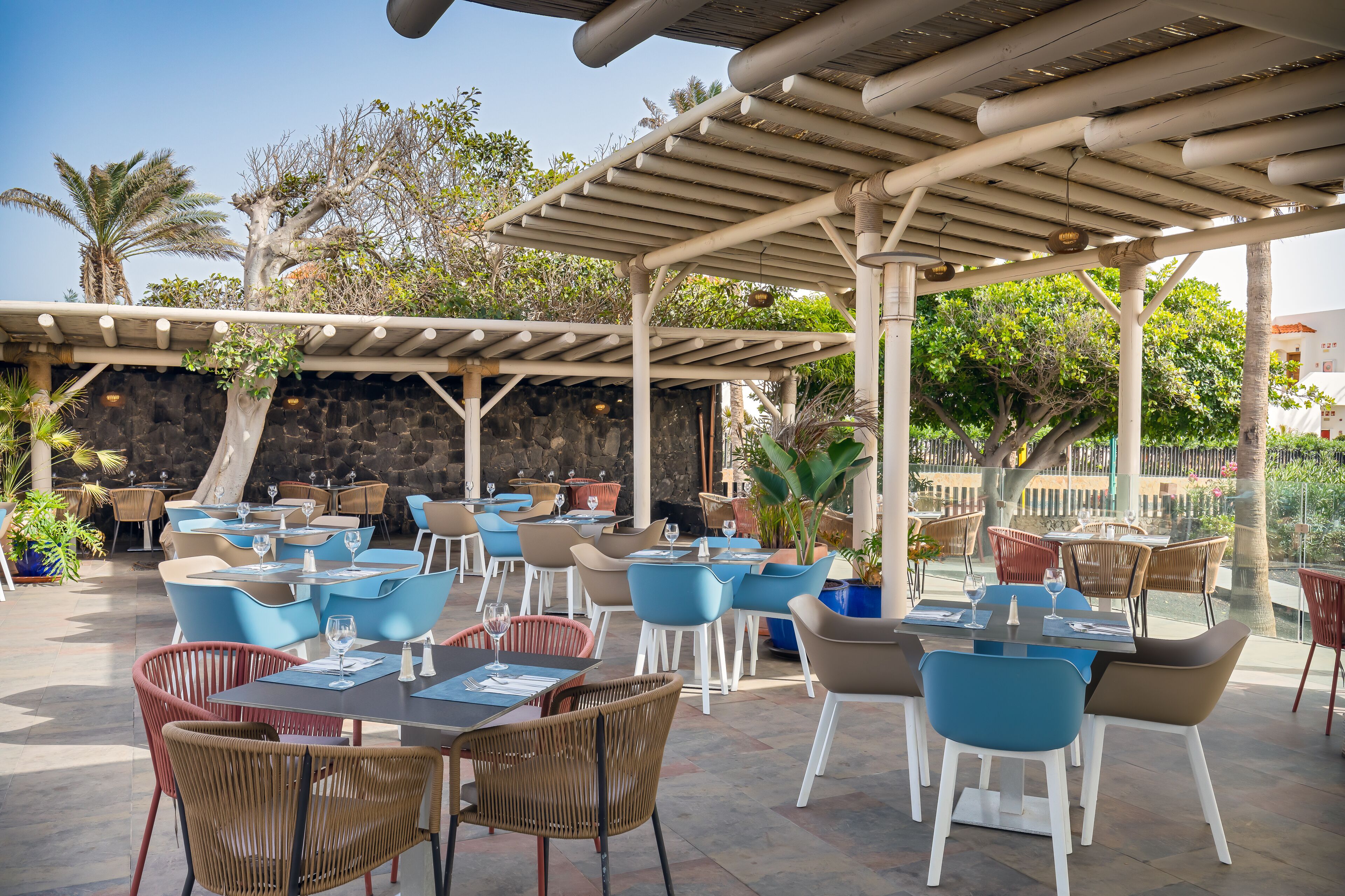 Un espace extérieur aménagé pour les repas, avec plusieurs tables et chaises, une structure en forme de pergola et un mur de pierre en arrière-plan.