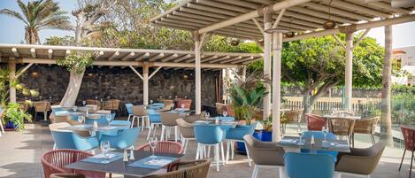 Un área de comedor al aire libre con varias mesas y sillas, una estructura de pérgola y una pared de piedra al fondo.