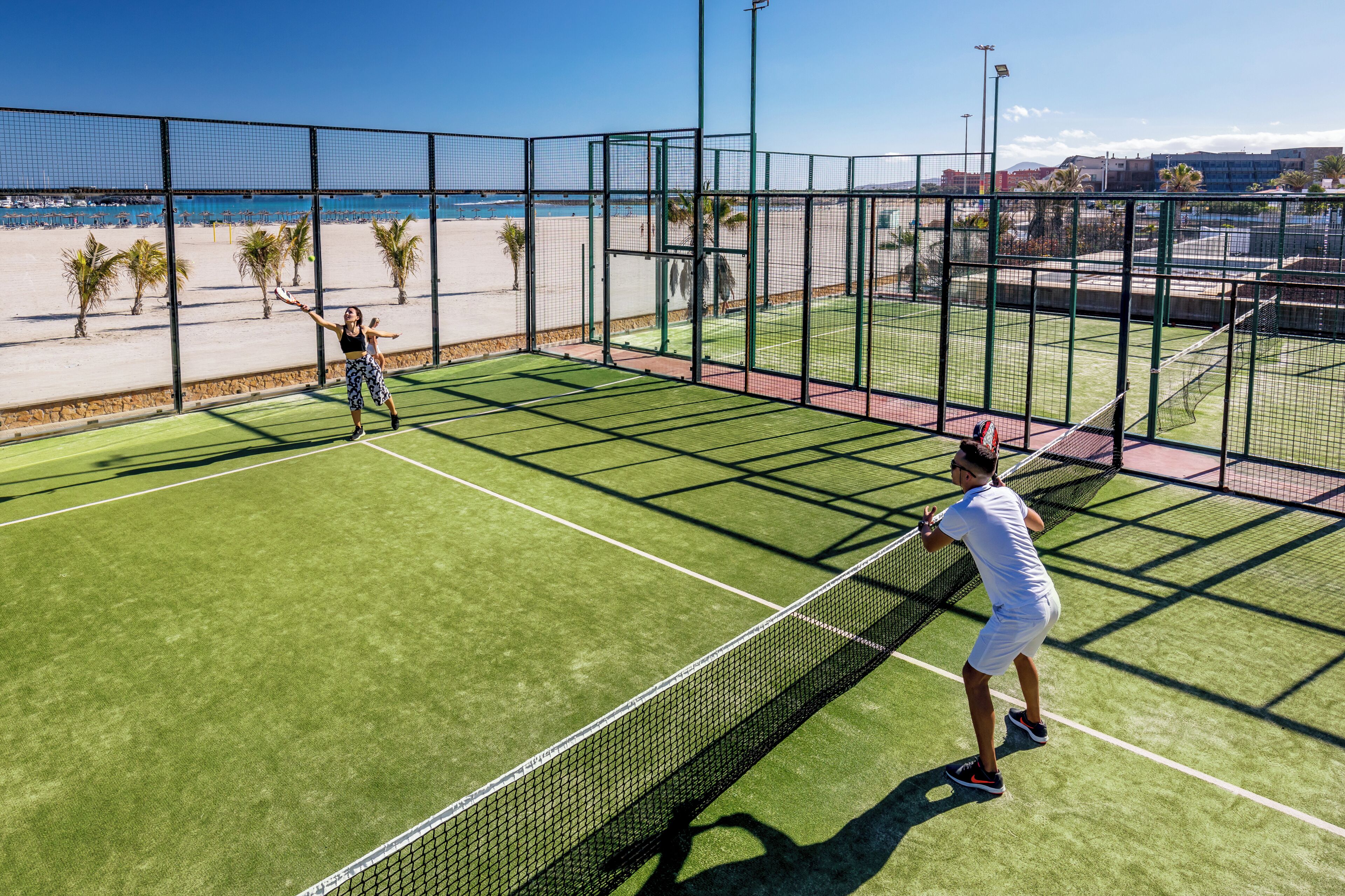 A tennis court with a net, surrounded by a fence, and a beach in the background.