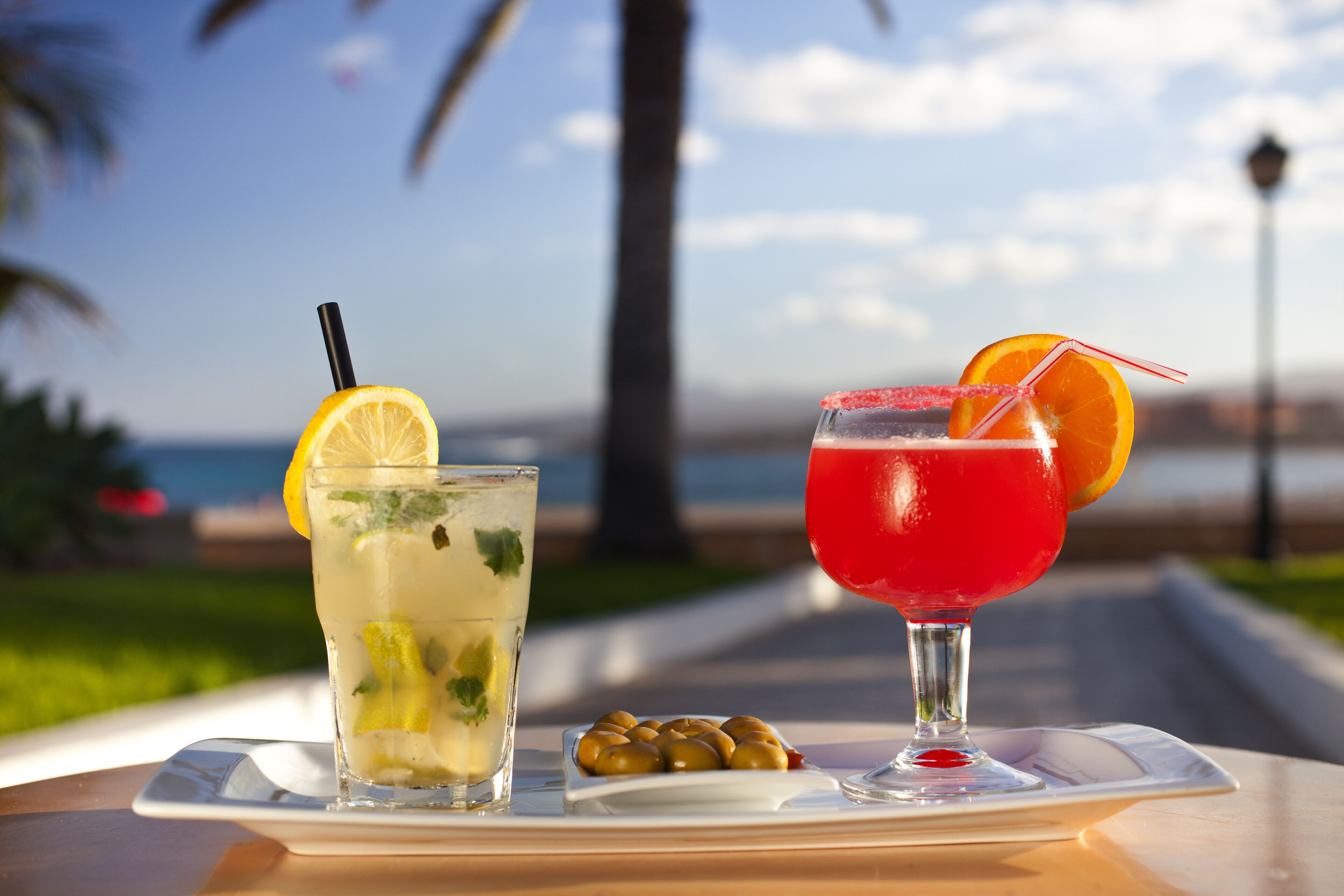 Two drinks on a tray with olives, set against a scenic background.