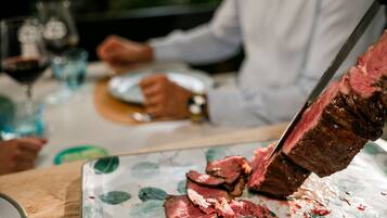 Une table sur laquelle on découpe une grande pièce de viande, des verres de vin rouge et une personne à l’arrière-plan.