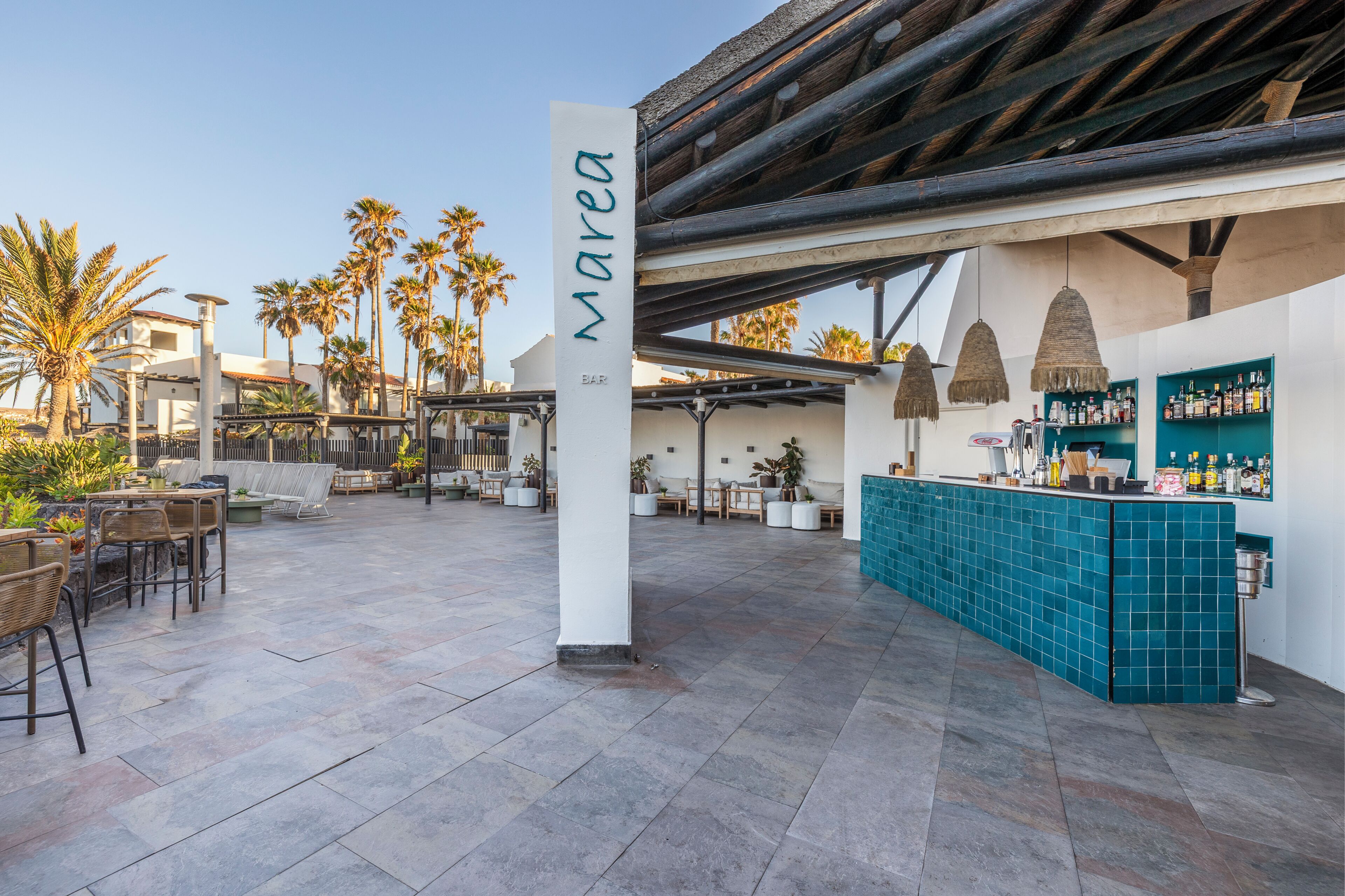 An outdoor bar area with a sign reading 'Marea BAR', seating arrangements, and a view of palm trees.