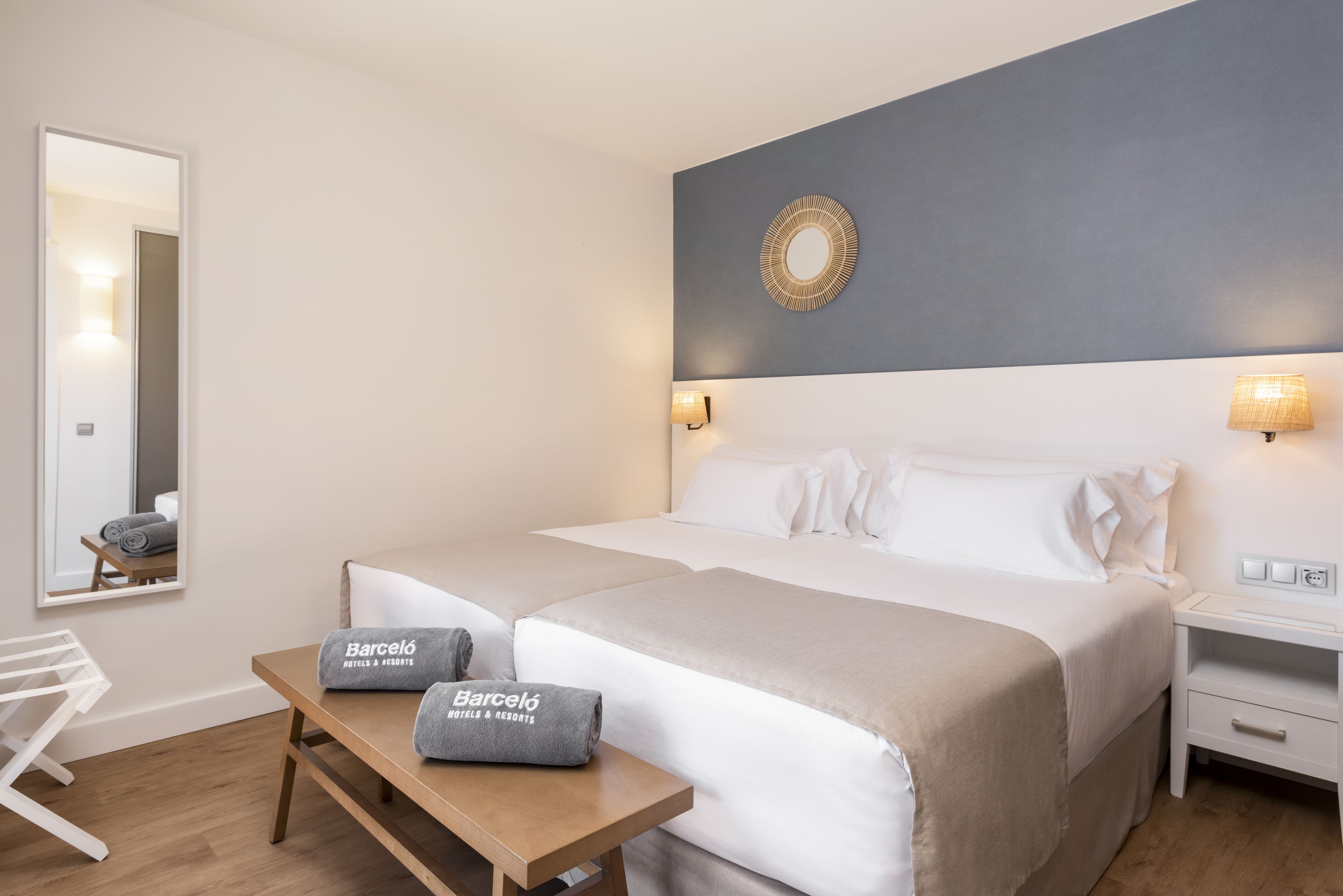 Hotelkamer met twee bedden, een houten tafel met handdoeken, een spiegel en een wandlamp.