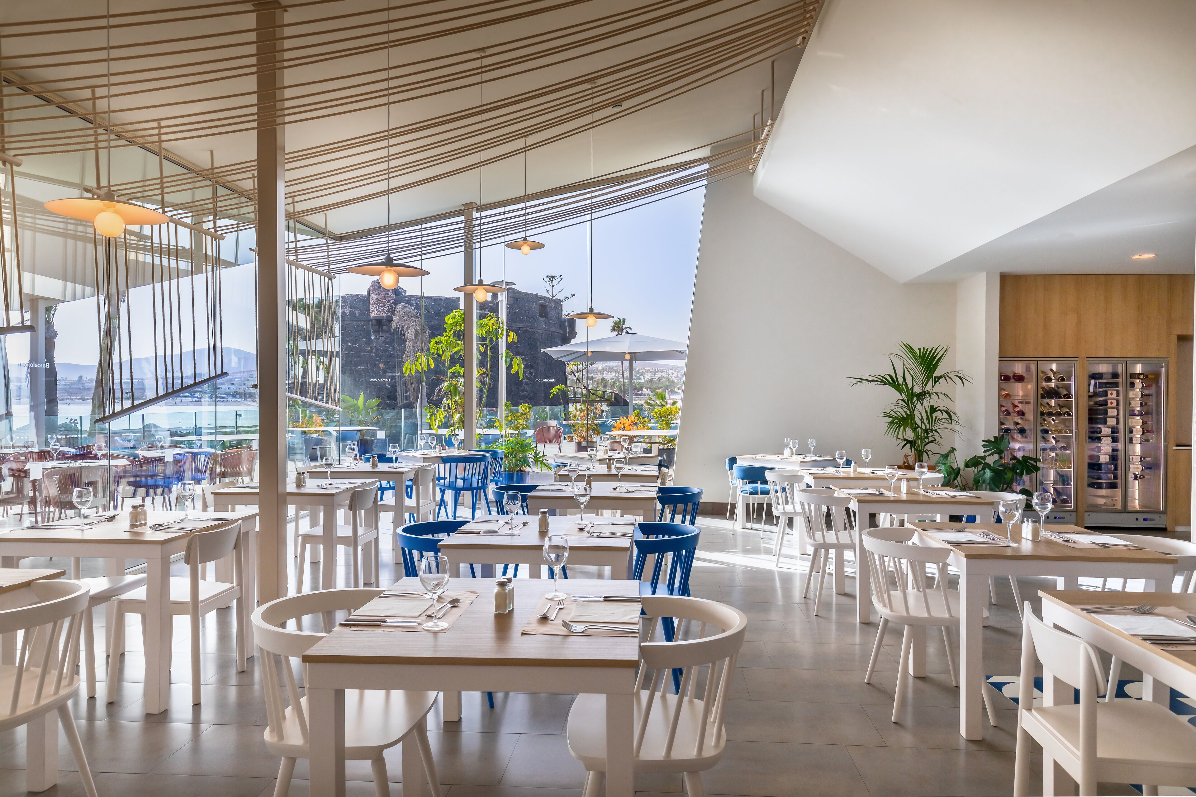 Ein modernes Restaurant mit weißen Tischen und blauen Stühlen, große Fenster mit Blick auf eine felsige Landschaft.